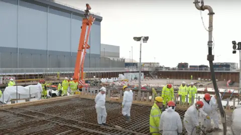 Sellafield Workers wearing white suits and florescent overalls as well as hard hats lay concrete and structural wiring at the treatment plant in Sellafield. There is a crane in the distance and other industrial buildings.