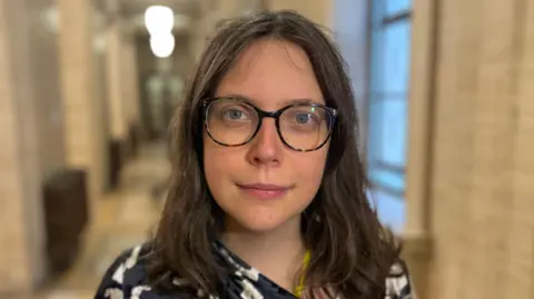 Dr. Viviane Gravey inside Stormont, she has long brown hair and is wearing dark framed glasses. Her top is black and white patterned.