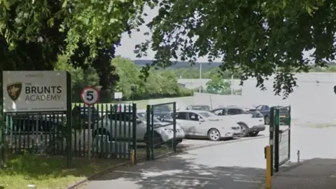 Google A general view of Brunts Academy Mansfield with cars parked behind gates and a sign reading The Brunts Academy