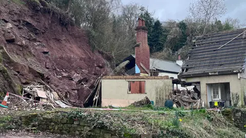 A steep rock wall that has suffered a landslide. Rubble from a partially collapsed house can be seen next to it.