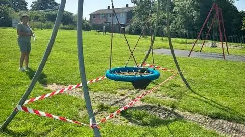 Liskeard Town Council A picture of a large swarm of bees on a blue basket swing which has been taped off by red and white tape. There is a woman in the background looking at swing. There is other play equipment in the background as well as houses. 