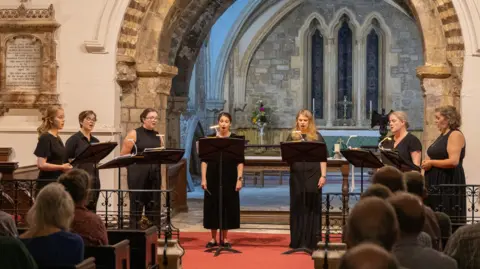 James O'Driscoll A performance inside Gloucester Cathedral by the Lady Clerks group. There are seven singers dressed in black stood in front of music stands with a number of people sat down watching.