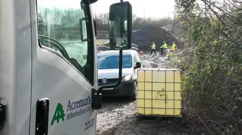 Arborist service vehicles parked at the site of the waste dump and there are workers in yellow high vis uniforms seen in the distance.