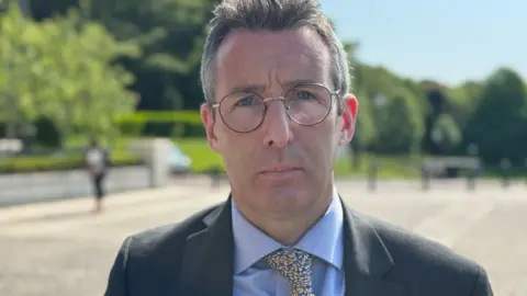 A man with dark hair and glasses, he is wearing a black blazer, blue shirt and blue and yellow tie