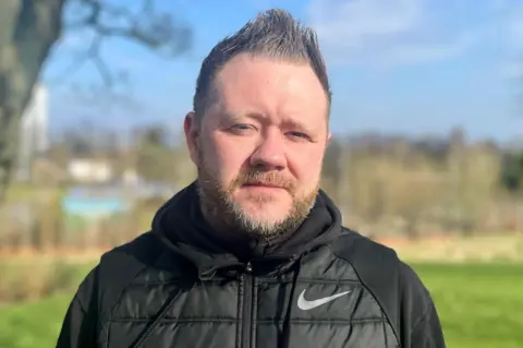 Head and shoulders shot of a bearded man in a black Nike tracksuit top