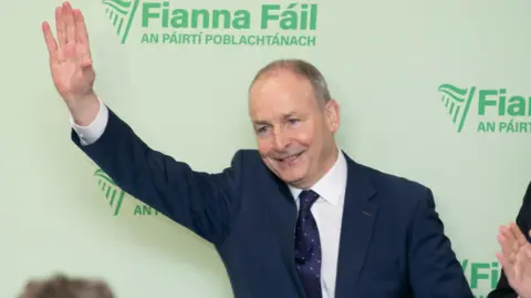 The Fianna Fáil leader, Micheál Martin, waving while at the ard fheis. He is standing in front of a green Fianna Fáil background