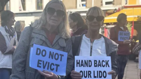 Jacqueline Jopson is on the left, she has long blonde hair and is holding a sign that says do not evict. Susan Rix is on the right and has short blonde hair. She is holding a sign that says Hand Back Our Lido. 