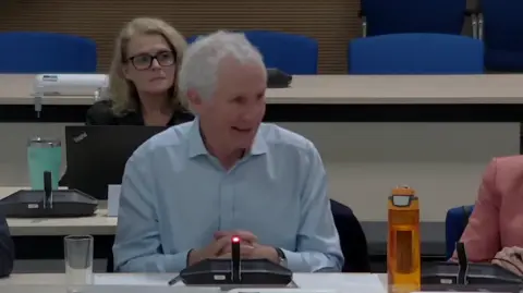 South Cambridgeshire District Council Peter Freeman speaking at a council meeting. He has short white hair and is wearing a light blue shirt, sat down on a table with a microphone in front of him.