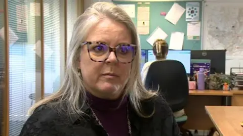 A woman with blonde hair past her shoulders looks at the camera, she's in an office room, behind her you can see someone sat at a desk looking at a computer. She has round glasses which are light brown with darker brown stripes, like an animal pattern around the rims. She has a dark top over a mauve jumper and wears a metal chain necklace.