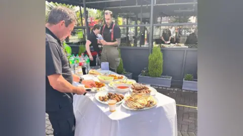 Nicky Brennan A man in a black t-shirt and trousers is standing next to a table full of food and drinks. A firefighter is holding a cup and standing on the opposite side of the table