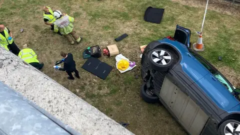 A blue car is on its side on the ground. Around it are people in high visibility vests. The car boot is open and there are shopping bags also on the ground as well as bits of the car's bumper. An air bag appears to have been deployed and can be seen through the drivers side window. 