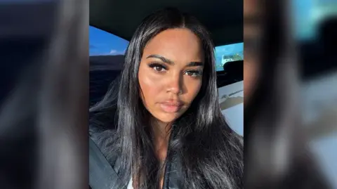 Klarisse Smith A woman with long straight dark hair sits in a car, wearing a black jacket and looking directly at the camera. Sunlight highlights her face, with a seatbelt visible across her chest and a blue sky outside the window.