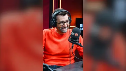 David Tennant smiles. He is wearing an orange jumper and wears black headphones. He sits in-front of a Radio 2 microphone.