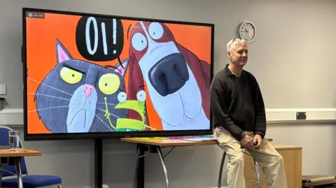 Jodie Halford A man sits on a table in front of a large screen with a cartoon picture of a cat, frog and dog with a speech bubble in black that says OI! The background of the cartoon is orange. 
