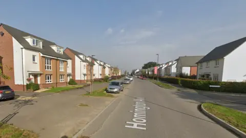 A Google street view image of a residential street. The properties are relatively new with some three-storey homes and some two-storey houses of differing sizes. There are some cars parked along the edges of the street and some grass and greenery dotted around.