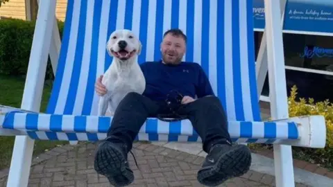 Paul Scott sits on an oversized, novelty blue and white striped deckchair, with a  white bulldog at his side. He is smiling and is wearing a blue top, black trousers and trainers.