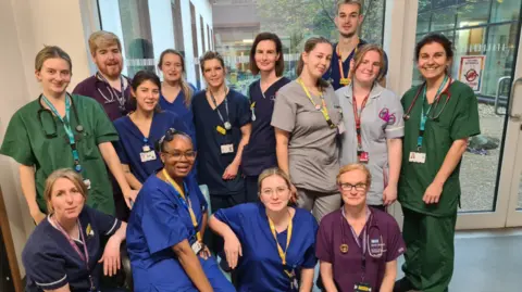 UHBW NHS A group of about a dozen clinical staff, wearing different coloured scrubs, standing in a hospital foyer with floor to ceiling glass windows behind. They are all smiling to camera.