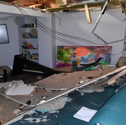 PA A living room interior wrecked by an explosion. The ceiling has partly collapsed, with large sections of plasterboard hanging down and exposed wooden roof timbers and wiring visible above. Debris, broken ceiling panels and rubble cover the floor and a sofa in the foreground. Built-in shelves of books and a framed picture remain on the walls, and a brightly coloured abstract painting leans against the far wall.