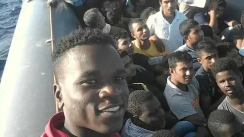 Andrew Kueth A number of young men crammed into a small boat with one person looking straight into the camera and smiling. There is a hint of blue water to the left of the picture.