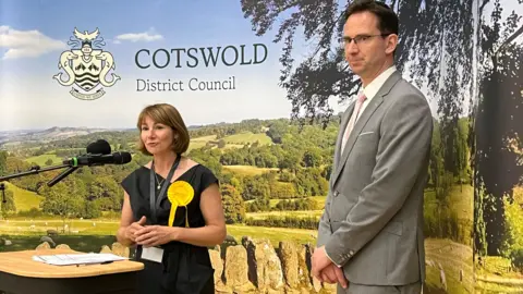 Councillor Spivey stands in front of a lectern as she delivers a speech. She is wearing a black dress with a yellow rosette on the chest.