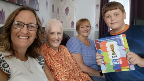 Contributor Four people sitting on a sofa in a living room from R to L Zac, a young boy, with brown hair wearing a blue and orange top holding his completed Panini FIFA sticker album. Next to him is his nan who has light brown short hair and wearing a blue short sleeve top. Next to her is an older lady with very short white hair and a long sleeved orange patterned blouse, next to her is a lady with shoulder length brown hair wearing a white short sleeved top, she is wearing glasses and a necklace. All four are smiling.