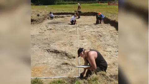 Six people measuring an archaeology trench on a sunny day