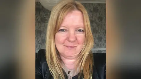 Maxine Hickman, with long blonde hair, wears a black leather jacket and a cream top underneath with gold buttons. She is smiling. She is in a room in a home decorated with grey flowery wallpaper. 