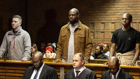 AFP via Getty Images The three of them standing. In front of them are three male court officials in black robes and ties