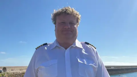 Bill Sadler stands by the harbour. He is wearing a crisp, white short‑sleeved shirt with shoulder epaulets. He has short curly light brown hair and is smiling.