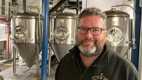 Chris France stands in front metallic brewery equipment and is smiling at the camera. He has short black/grey hair and a beard and is wearing glasses.
