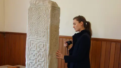 Rachel Pickering A woman examines a patterned stone pillar. She has a camera in one hand, dark hair in a ponytail and glasses on top of her head.