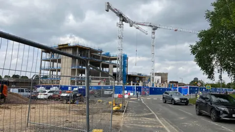 Two tall cranes dominate the skyline at the Royal Shrewsbury Hospital. Metal barriers are on the left, through which can be seen piles of sand and aggregate, as well as a car park beyond. Two cars are leaving the hospital site on the right, with further blue hoardings behind them. A partially built concrete building is next to the cranes, with exposed sides showing four storeys