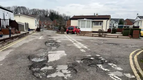 A junction of roads on a mobile home park. There are some large pits in the foreground, and are pulled away in distance. A cream colored mobile with a brown roof and front porch sits on the curve of the house junction, with a small red car stands in front. Double yellow lines are running on the edges of the road. Other mobiles are visible on the left hand side and some gates are open in the background, with red brick houses. 