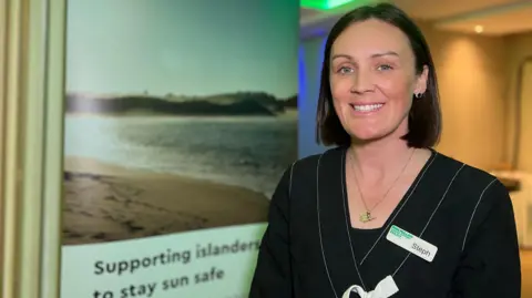 Steph is wearing a black outfit and name badge stands indoors beside a display banner showing a beach scene and the words “Supporting islanders to stay sun safe,” in a softly lit conference or event space.