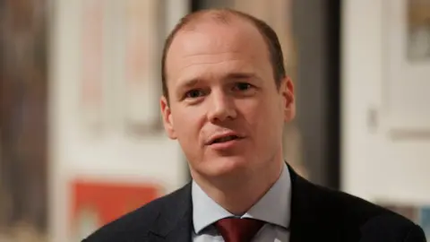 PA Media Gordon Lyons wears a dark suit with a light shirt and burgundy tie. He has short brown hair.  He is looking into the camera.