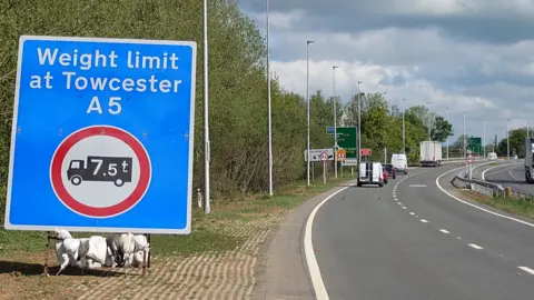 National Highways A large blue traffic sign weighed down by sandbags. It reads 'Weight limit at Towcester A5' and shows a red and white symbol of an HGV with '7.5 tonnes' on it. The sign is beside a dual carriageway where light traffic is shown moving freely beneath leaden skies.