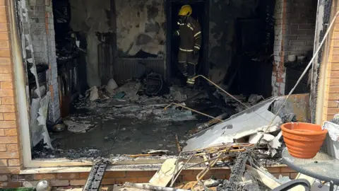 Derbyshire Fire and Rescue Service Debris inside and outside one of the rooms at the home. A firefighter in full protective gear in stood inside.