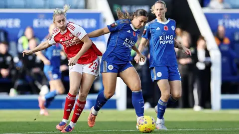 Getty Images A shot from Leicester ladies vs. Arsenal shows an Leicester player in a blue strip on the ball and fending off a challenge from an opposing player, in red. A second Leicester team-mate looks on as the possessing player dribbles ahead.