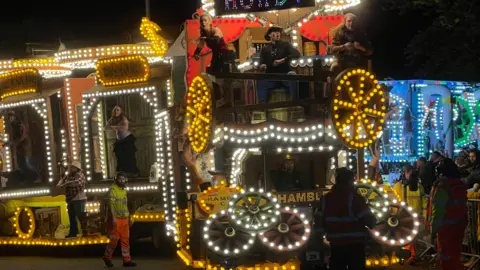 An illuminated carnival cart. There are a number of performers on it looking out towards the crowds. The cart has a number of illuminated wheels on it and appears to have a Western theme.