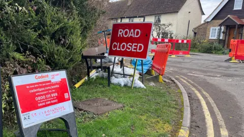 Tony Fisher/BBC A "Road Closed" sign on a grass verge by the road. There is another sign next to it which has details for Cadent Gas. There are plastic hoardings on the road and a wooden bench is also on the grass verge.