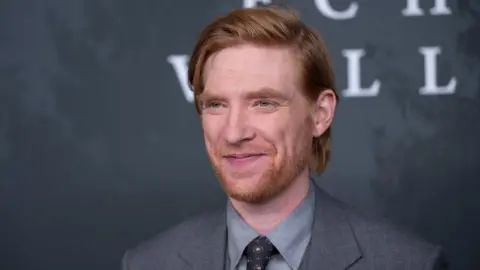 Getty Images Actor Domhnall Gleeson wearing a grey suit jacket, light grey dress shirt, and patterned tie stands in front of a dark backdrop with large, partially visible white letters. He has ginger hair styled neatly, and the background features a textured, mottled design.