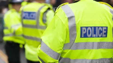 Three police officers standing outside with their backs to the camera. They are all wearing yellow fluorescent jackets with grey stripes along the back and arms. On the back of each jacket is a blue rectangle with the word, POLICE inside.