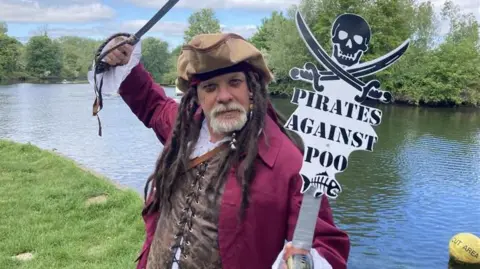 Darren poses in his pirate clothing holding a cutlass over his head and a sign saying "Pirates against Poo", which has a skull and crossed cutlasses at the top. He wears a maroon jacket, laced white shirt and brown tunic, and stands by the side of a river.