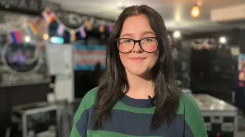 Hermione smiles and looks into the camera. She is wearing glasses with a green and blue jumper.
