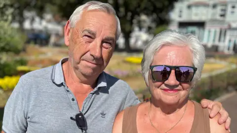 John and Sue Wackett standing by the seafront in Clacton. They both have grey hair. Sue is wearing a brown vest top and sunglasses. John is wearing a grey polo top and squinting due to the sun.