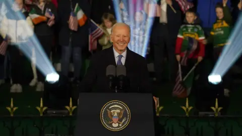 A man with white hair smiles. He is standing at a podium with an American eagle design on the front. He is wearing a suit. Behind him is a crowd, with a mixture of Irish and American flags. 
