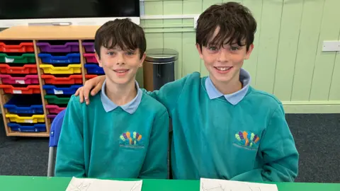 Twins Jakey and Sammy, who both have short, dark hair, sit behind a green desk in their classroom. Sammy has his arm around Jakey and they are wearing green school sweaters and light blue polo necks