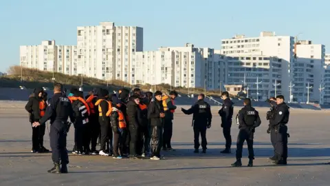 PA Media People thought to be migrants are spoken to by police officers after failing to board a small boat off the coast of Dunkirk, France. They are standing on a beach with tower blocks behind them.
