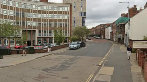 A city centre road, with town houses to the right and an office block to the left, extends away from the camera. It is cloudy above.