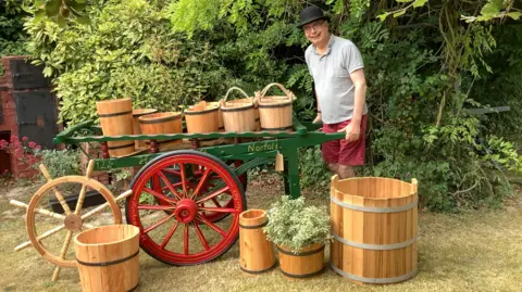 Alan Paulus Alan Paulus wearing shorts, a T-shirt and a black bowler hat holding a large green cart, which has red wheels. On top and around the cart are buckets of all sizes.